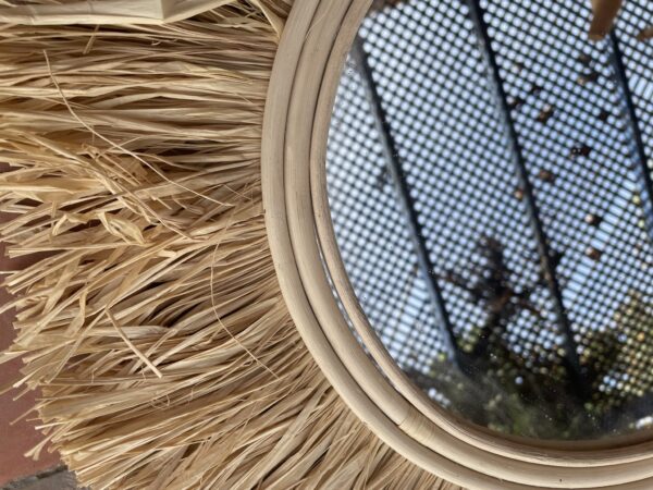 Photographie d'un miroir rond avec un contour en osier et franges en raphia. Diamètre 50 cm.